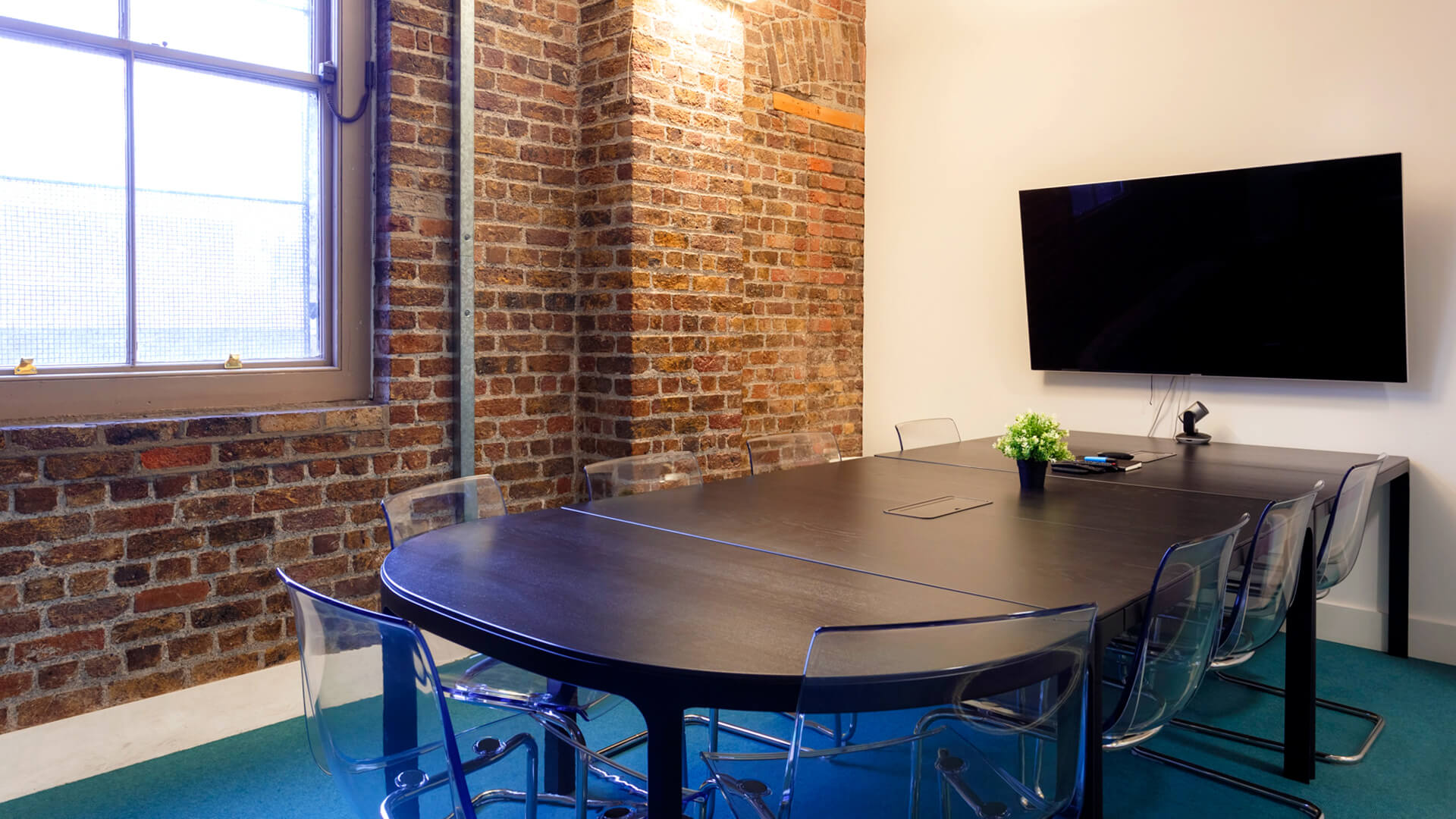Chairs are arranged around a wooden desk and display screen in a meeting room with exposed brick walls