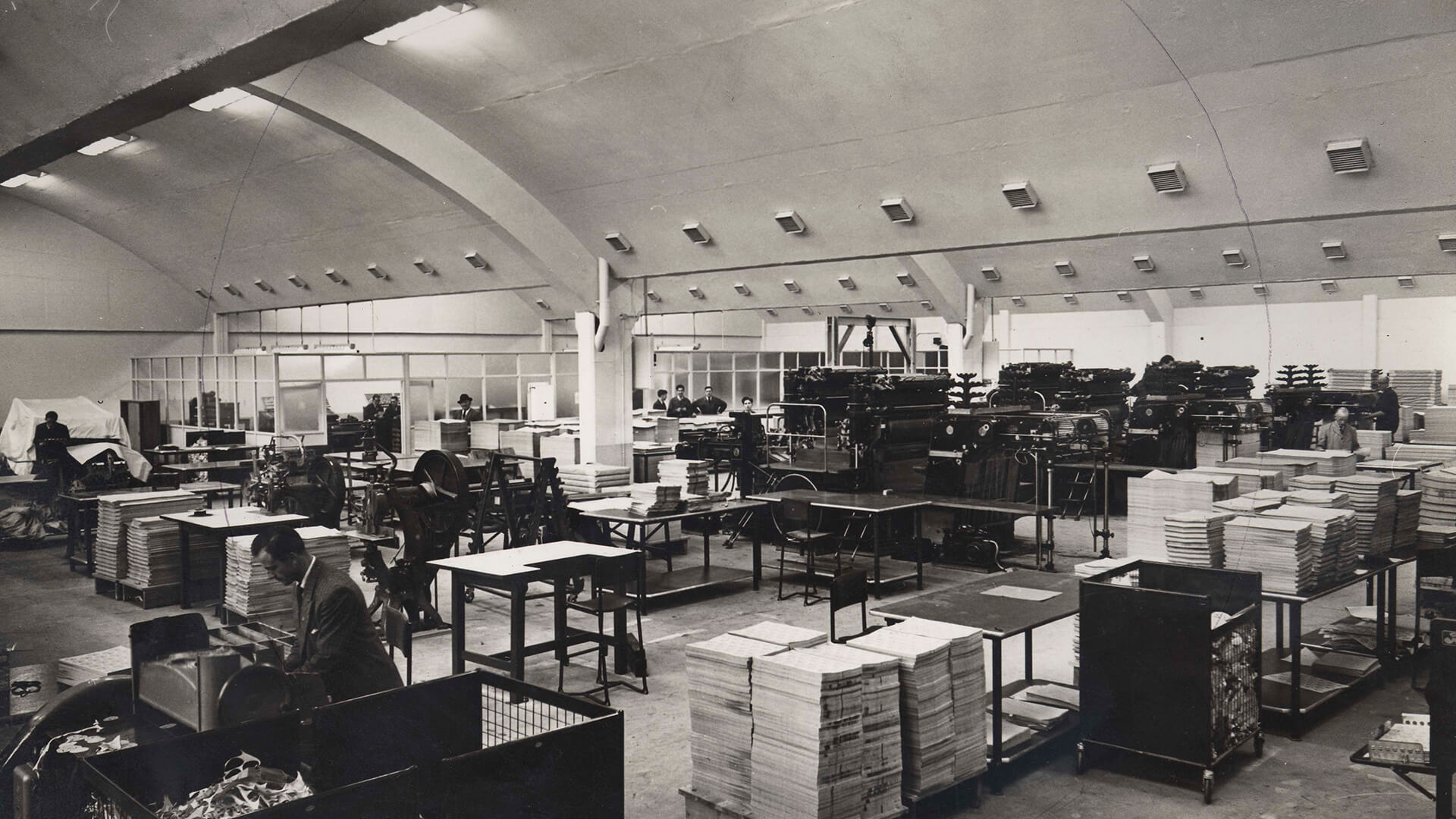 A black-and-white historical shot of the Guinness Printworks interior showing stacks of paper and machinery in a large, open space