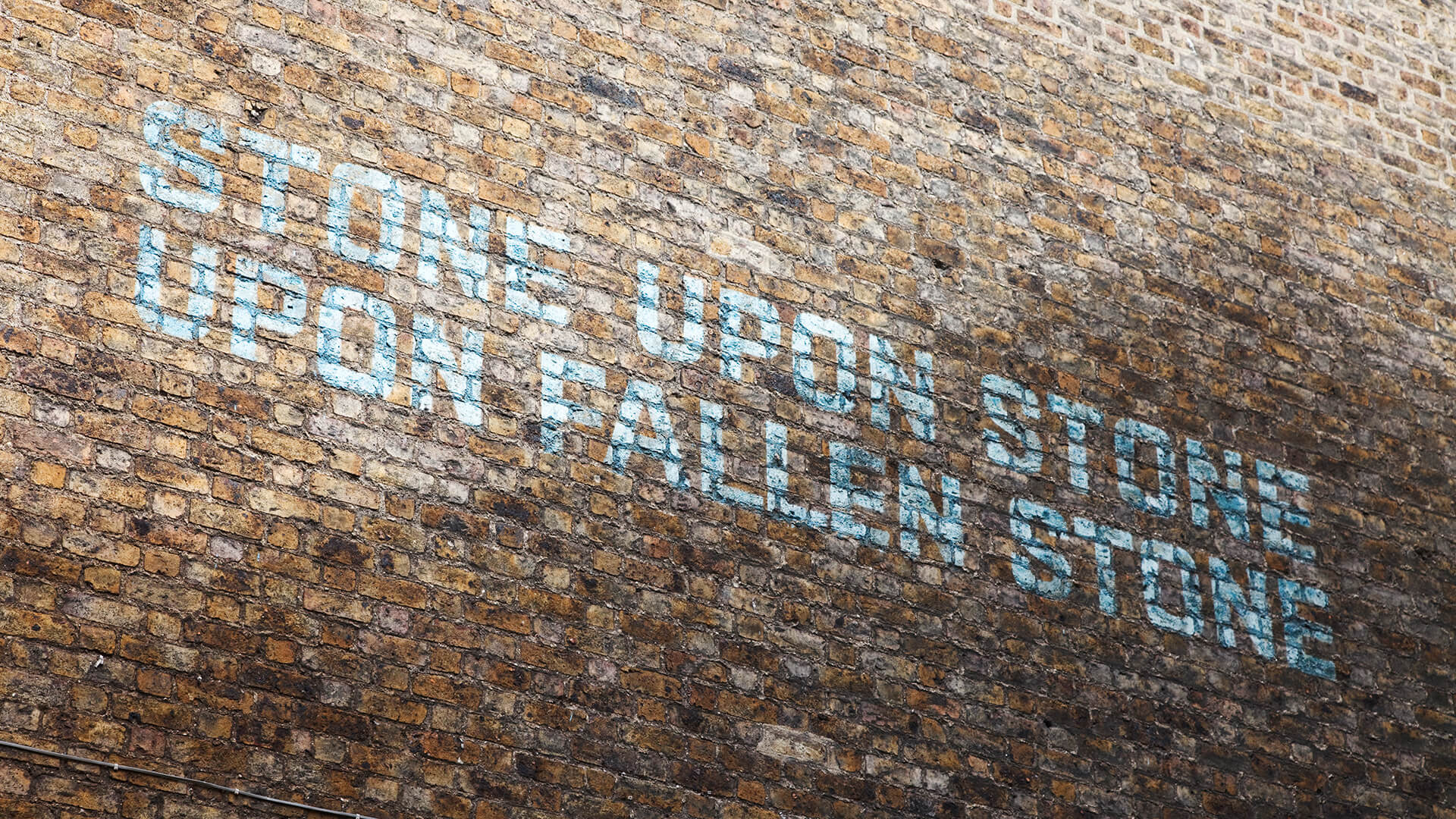Uppercase text is painted in pale blue on the brick wall of the Guinness Vathouse. The text reads: "Stone Upon Stone Upon Fallen Stone"
