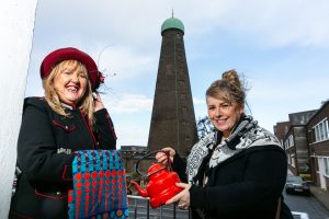 Two smiling women stand in front of St Patrick's tower holding colourful fabric and a red teapot